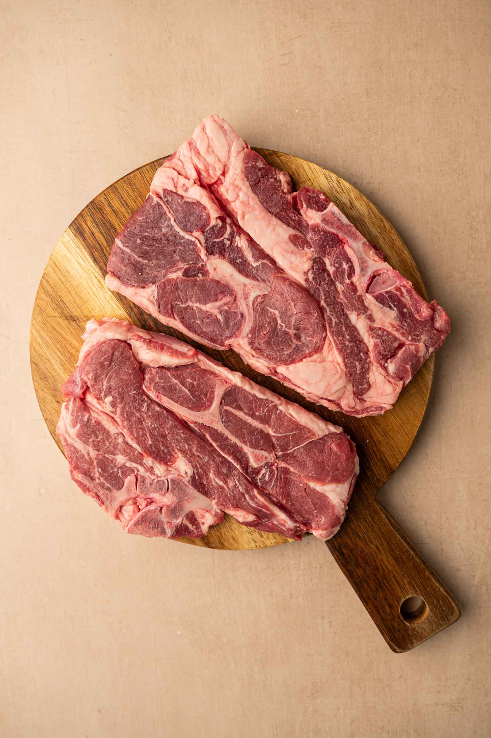Two raw lamb shoulder chops on a round wooden cutting board with a handle, placed on a light-colored surface.