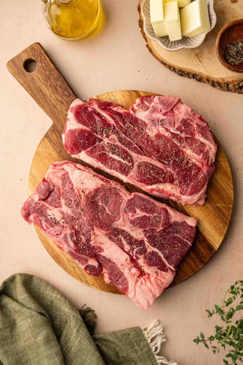 Two raw, seasoned shoulder chops on a wooden cutting board, with butter, pepper, olive oil, and a green cloth nearby.
