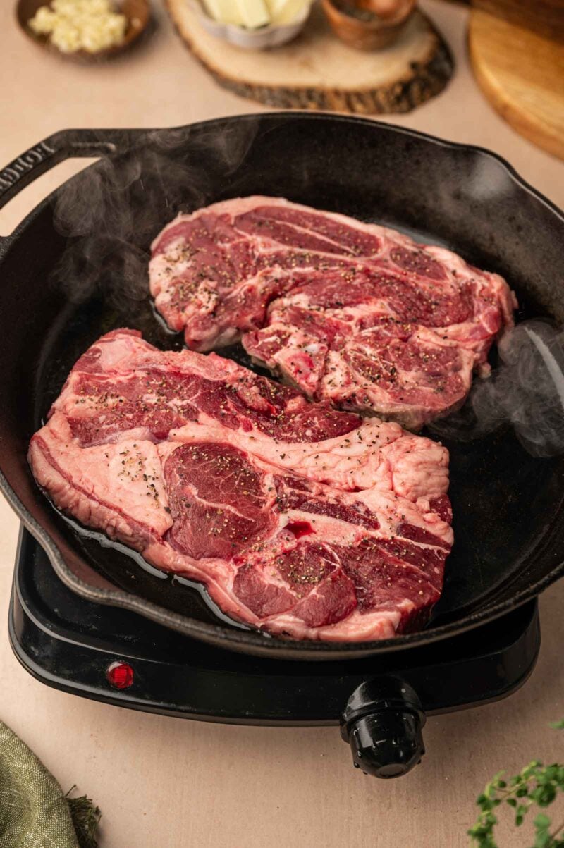 Two raw, seasoned lamb shoulder chops are cooking in a black cast iron skillet on an electric stovetop. Steam is visible around the meat.