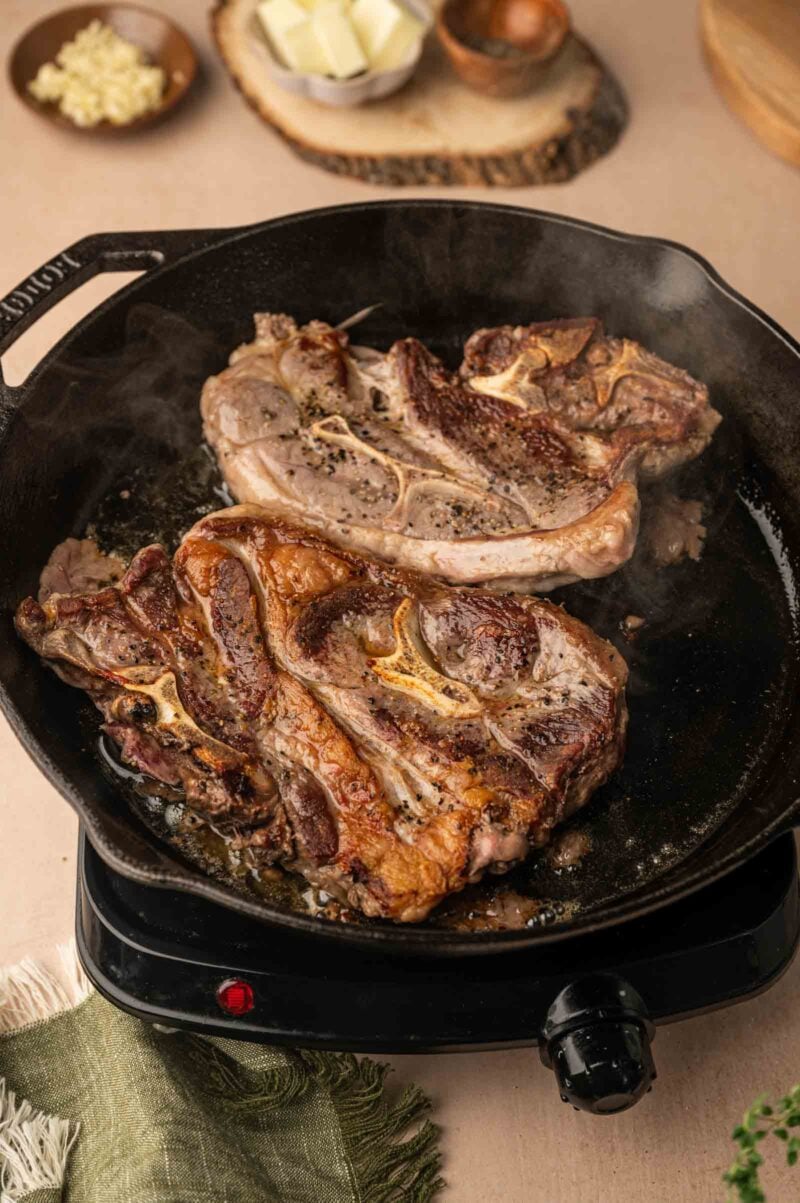 Two bone-in lamb shoulder chops cooking in a cast iron skillet on an electric stove, with seasonings and butter in the background.
