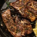 Two cooked lamb shoulder chops seasoned with herbs and garlic are shown in a black cast iron skillet with lemon wedges on the side.