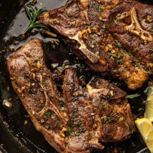 Two cooked lamb shoulder chops seasoned with herbs and garlic are shown in a black cast iron skillet with lemon wedges on the side.