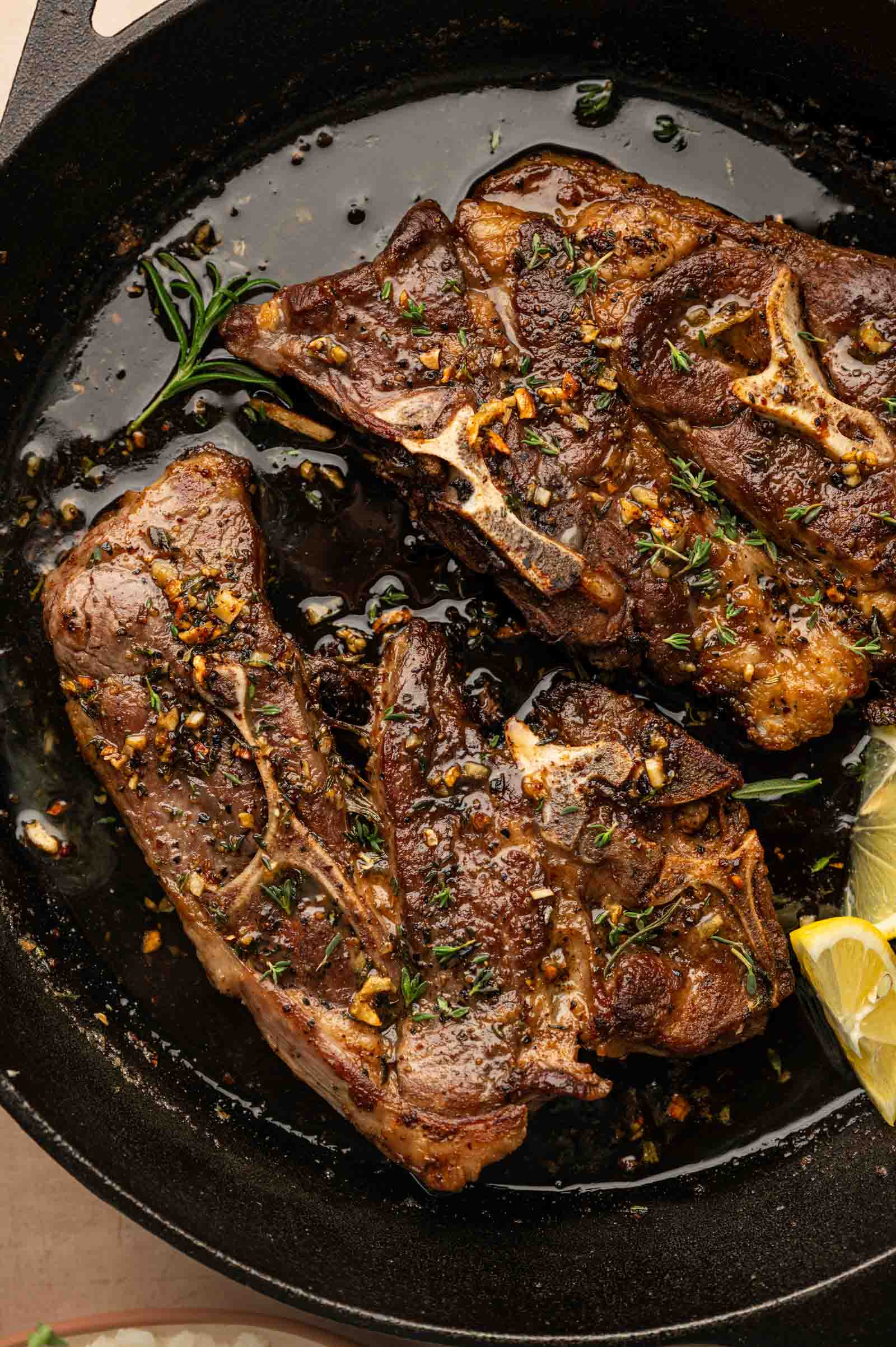 Two cooked lamb shoulder chops seasoned with herbs and garlic are shown in a black cast iron skillet with lemon wedges on the side.