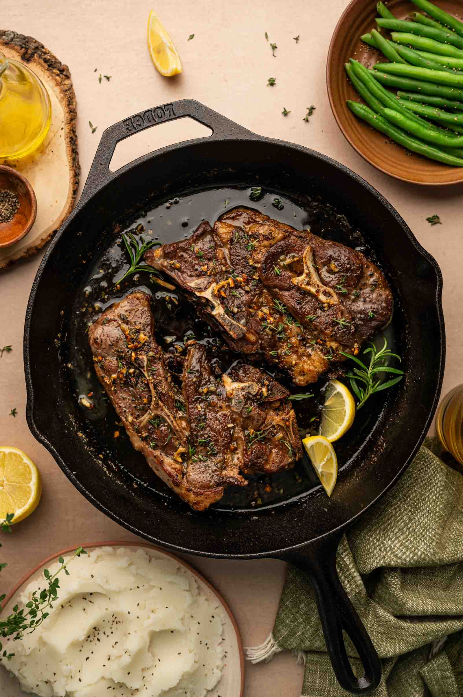 A cast iron skillet with two cooked lamb shoulder chops, garnished with herbs and lemon, surrounded by mashed potatoes and green beans on a table.