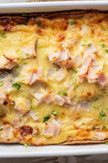 Baked casserole topped with melted cheese and pieces of ham or turkey in a white rectangular dish, placed on a woven mat.
