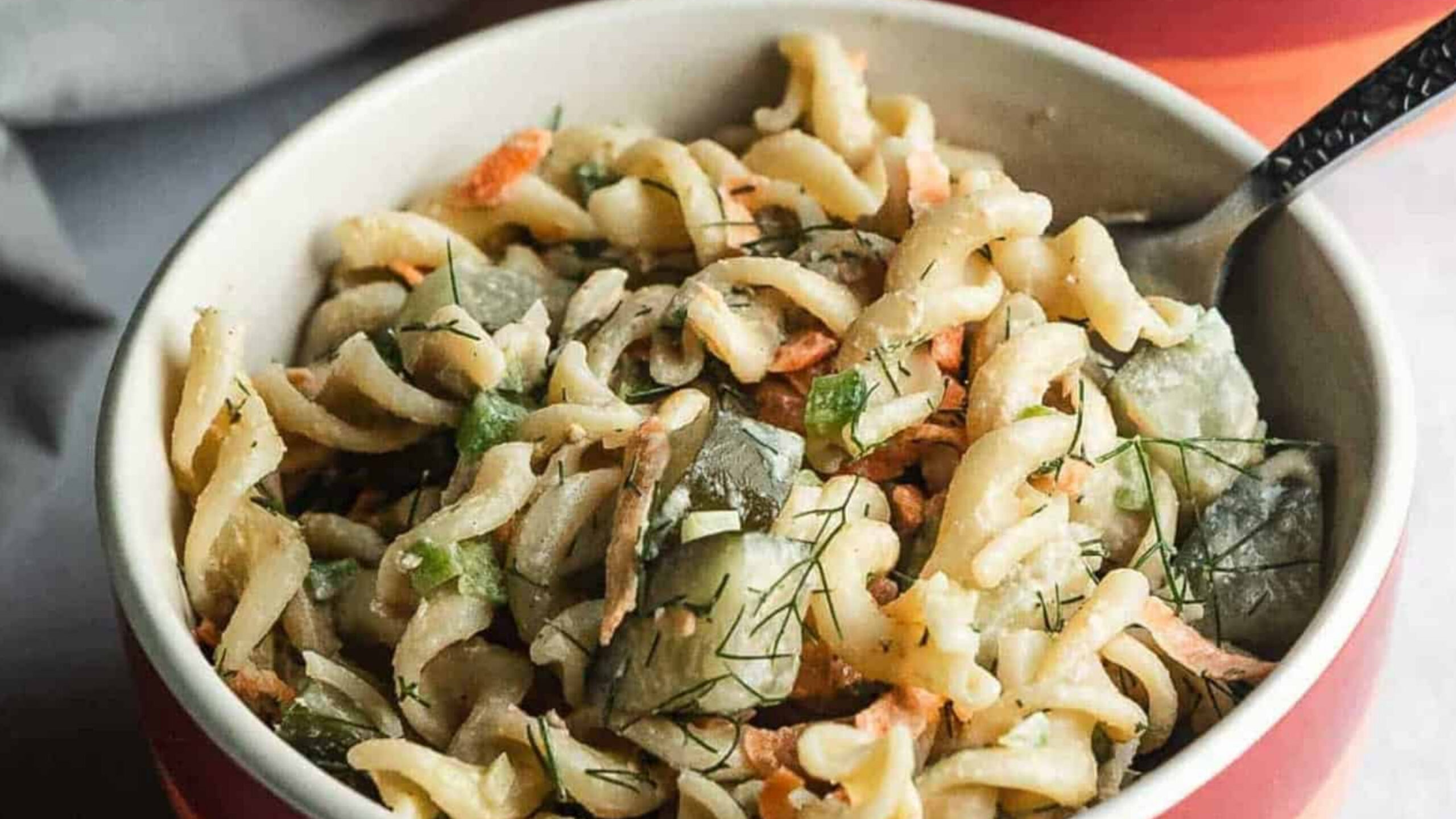 A bowl of rotini pasta salad with cucumbers, carrots, and herbs, with a fork inside.