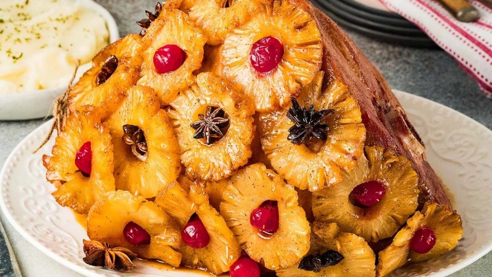 A baked ham decorated with pineapple rings, maraschino cherries, and star anise, served on a white plate. Mashed potatoes are visible in the background.