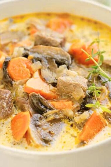 A bowl of creamy soup containing chunks of beef, carrots, mushrooms, and herbs, with a spoon and a black-and-white napkin nearby.
