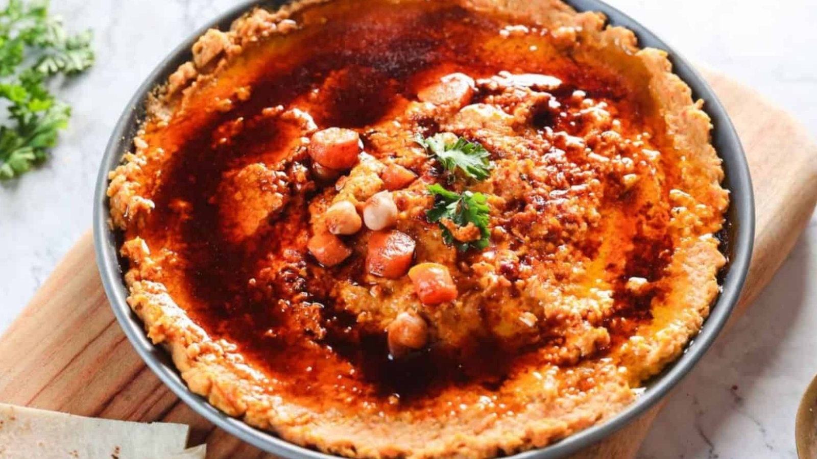 A bowl of hummus topped with spicy red sauce and pieces of chickpeas and carrots, garnished with parsley. The bowl is placed on a wooden board with a partially visible sprig of herbs and flatbread nearby.