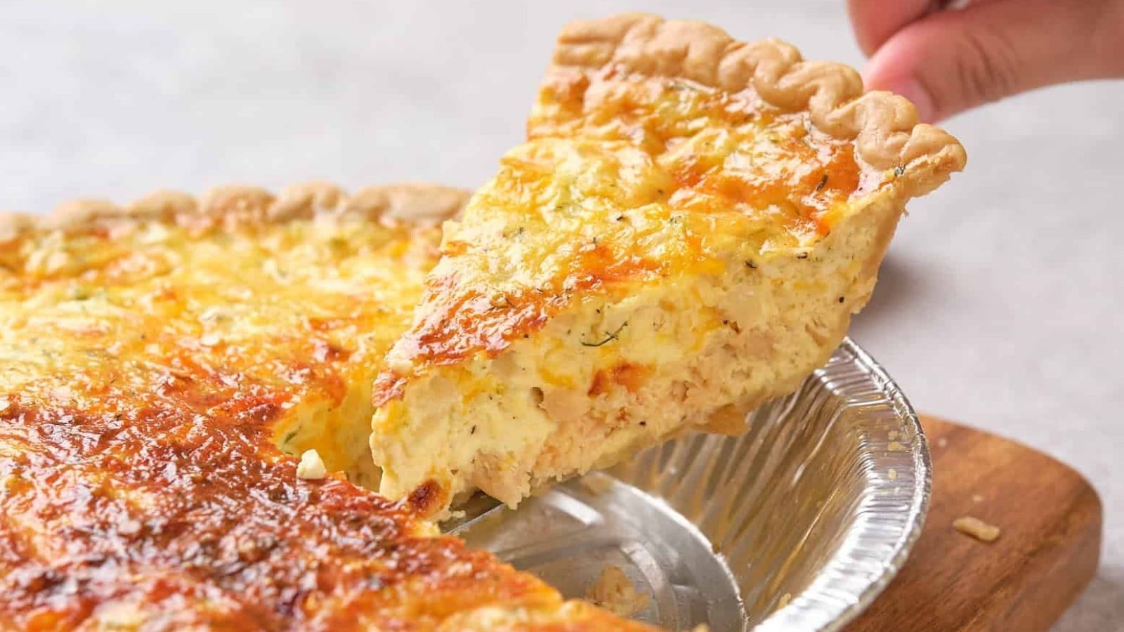 A hand lifting a slice of salmon quiche from a pie tin, revealing a golden, crispy top and creamy filling.