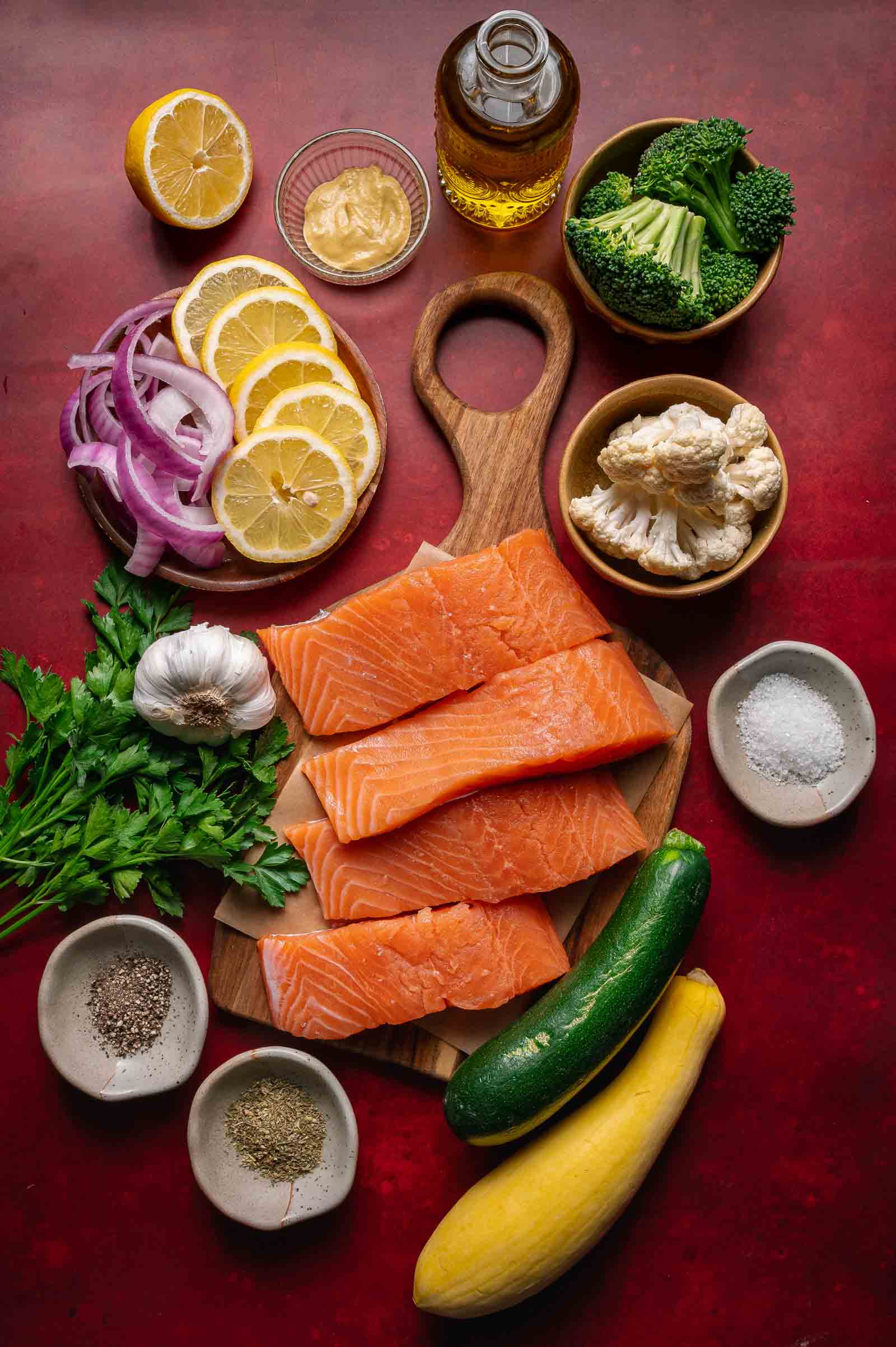 Raw salmon fillets on a wooden board surrounded by sliced lemon, red onion, garlic, parsley, zucchini, yellow squash, broccoli, cauliflower, olive oil, mustard, salt, and pepper.