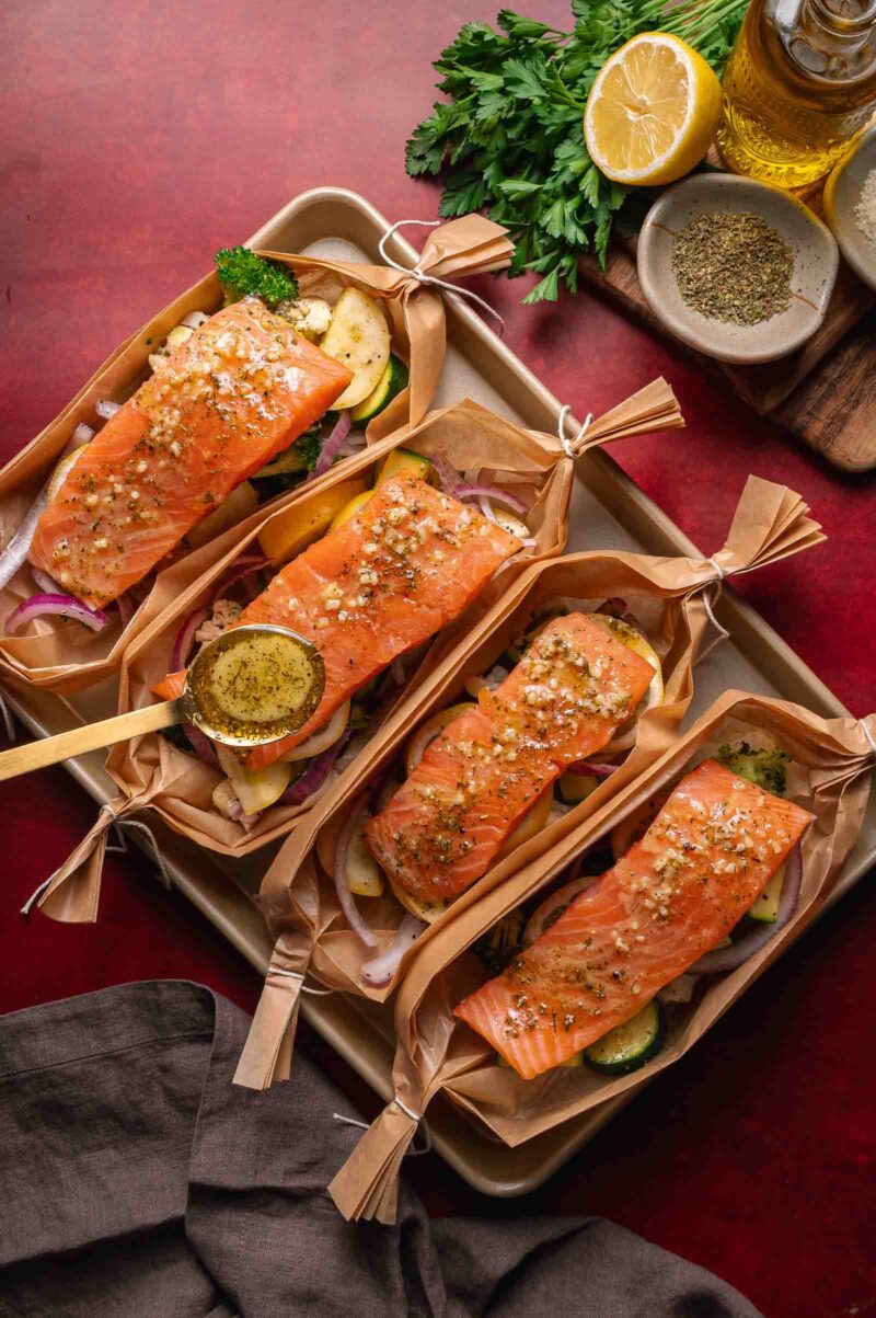 Four parchment boats filled with seasoned salmon fillets and vegetables on a baking tray, with a spoon drizzling sauce and fresh herbs, lemon, and spices nearby.