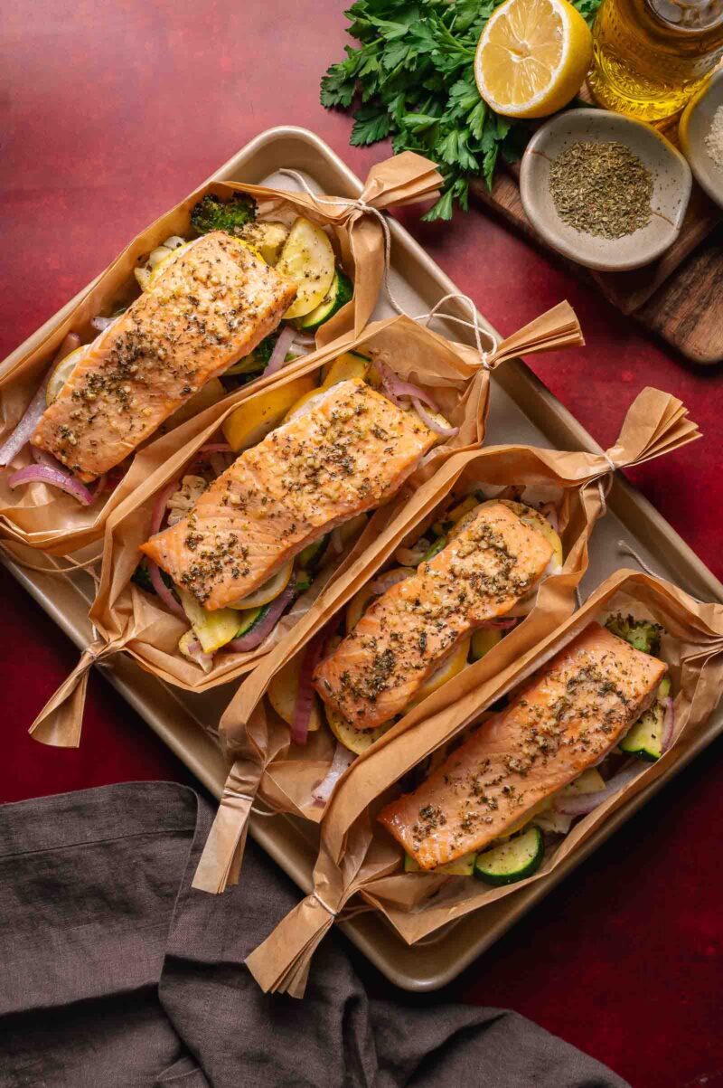 Four baked salmon fillets, seasoned with herbs, are placed on sliced vegetables and wrapped in parchment paper boats on a baking tray. Fresh parsley, lemon, and seasonings are nearby.