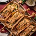 Four salmon fillets seasoned with herbs are placed on top of mixed vegetables and wrapped in parchment paper boats, displayed on a baking tray with utensils and ingredients nearby.