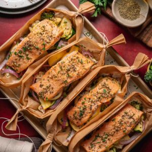 Four salmon fillets seasoned with herbs are placed on top of mixed vegetables and wrapped in parchment paper boats, displayed on a baking tray with utensils and ingredients nearby.