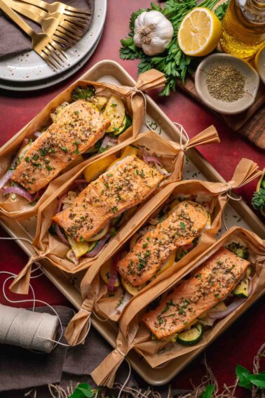 Four salmon fillets seasoned with herbs are placed on top of mixed vegetables and wrapped in parchment paper boats, displayed on a baking tray with utensils and ingredients nearby.