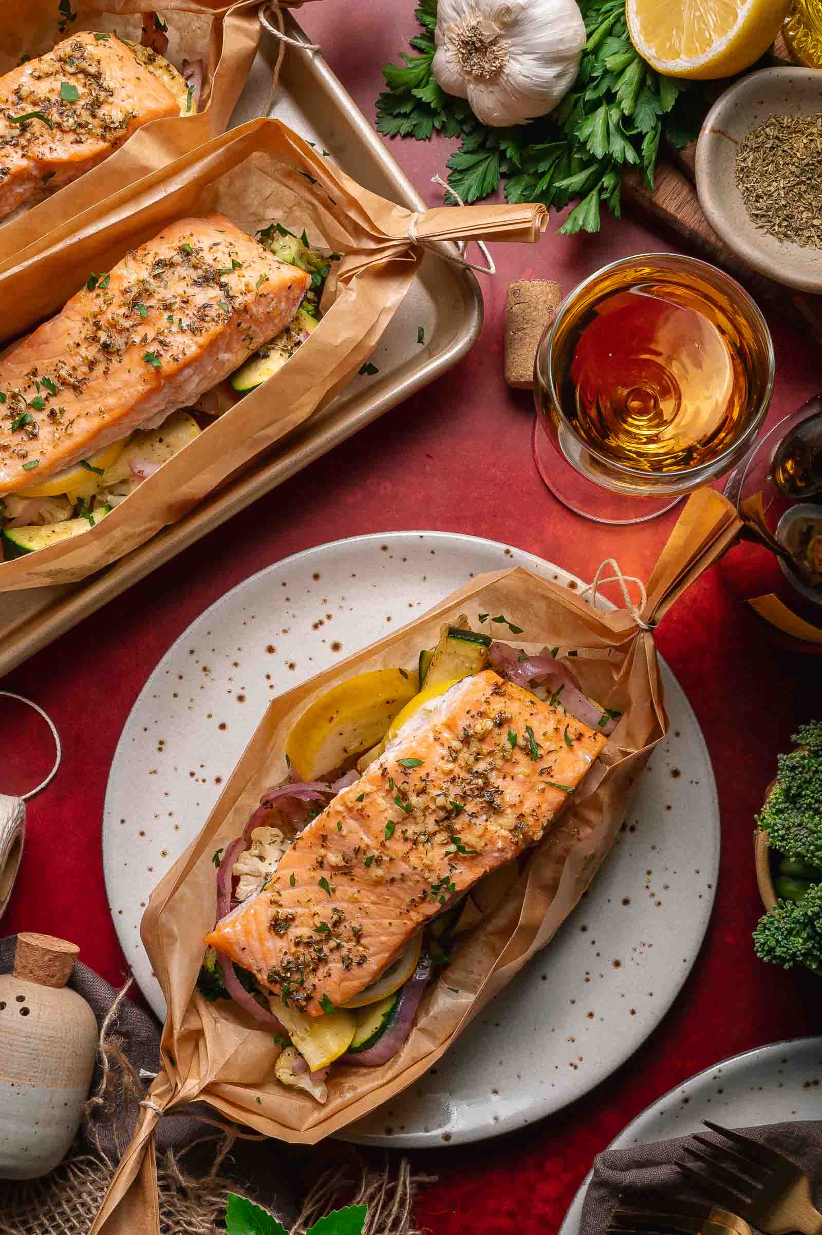 Baked salmon fillet in a parchment paper boat atop sliced vegetables, served on a plate with herbs, lemon, and a glass of amber liquid nearby.
