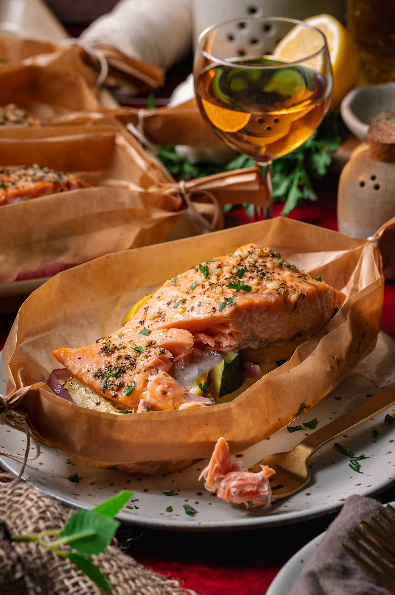 A baked salmon fillet with herbs and vegetables served in parchment paper on a plate, with a fork and a glass of white wine nearby.
