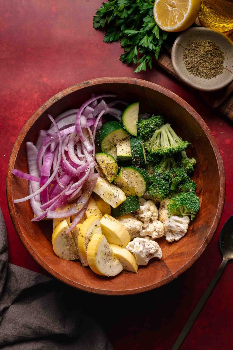 A wooden bowl containing sliced red onions, yellow squash, zucchini, broccoli, and cauliflower, seasoned with black pepper, sits on a red surface near herbs, a lemon, and spices.
