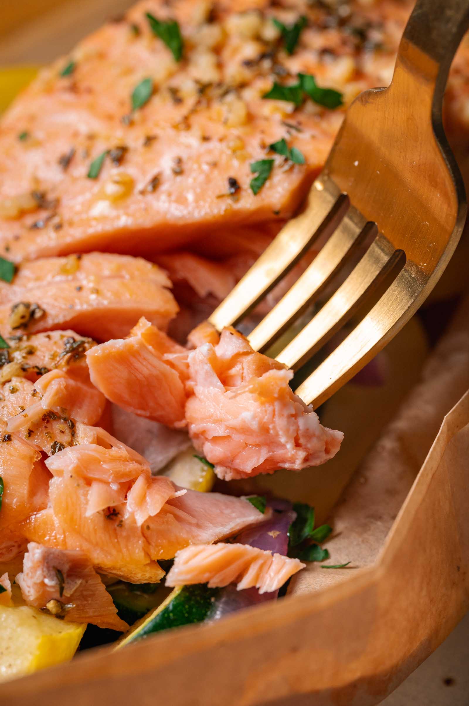 A close-up of a fork flaking a piece of cooked salmon garnished with herbs, served on a bed of vegetables.
