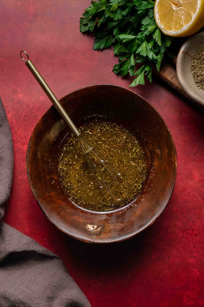 A wooden bowl containing a whisk and a mixture of oil and herbs, with parsley, half a lemon, and a dish of dried herbs nearby on a red surface.