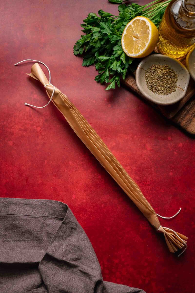 Folded parchment tied at both ends, placed on a red surface near fresh parsley, half a lemon, dried herbs, olive oil, and a brown cloth.
