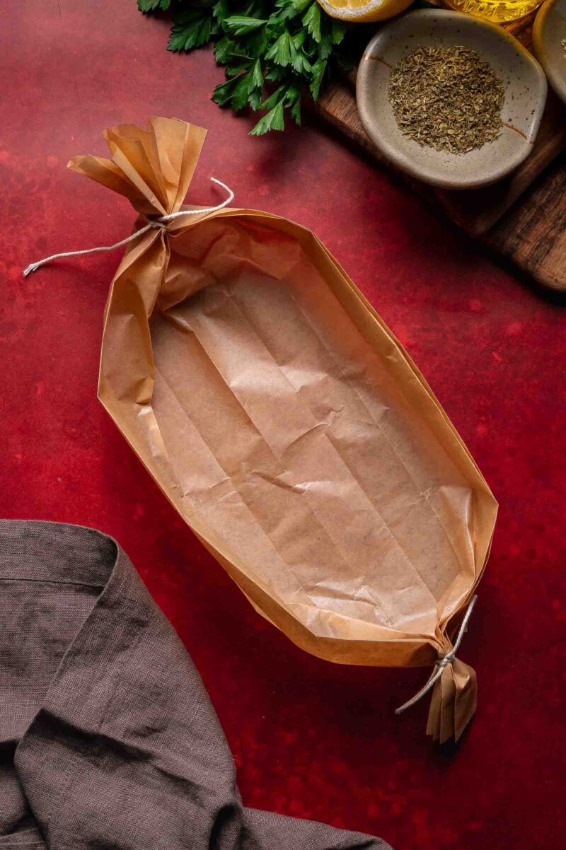 An empty parchment paper boat tied at both ends sits on a red surface near a gray napkin, herbs, and small bowls of spices.