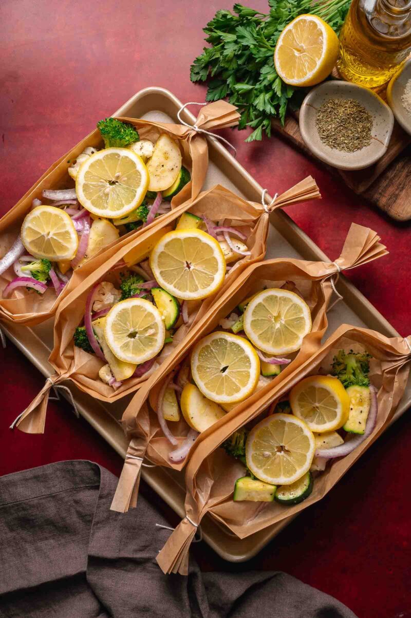 Three parchment paper boats filled with sliced lemon, zucchini, onions, and herbs are arranged on a baking tray, with fresh parsley, spices, and olive oil nearby.