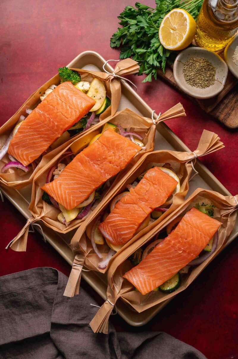 Four raw salmon fillets are placed on top of sliced vegetables in parchment paper boats, ready for cooking, on a baking tray with herbs, lemon, and spices nearby.