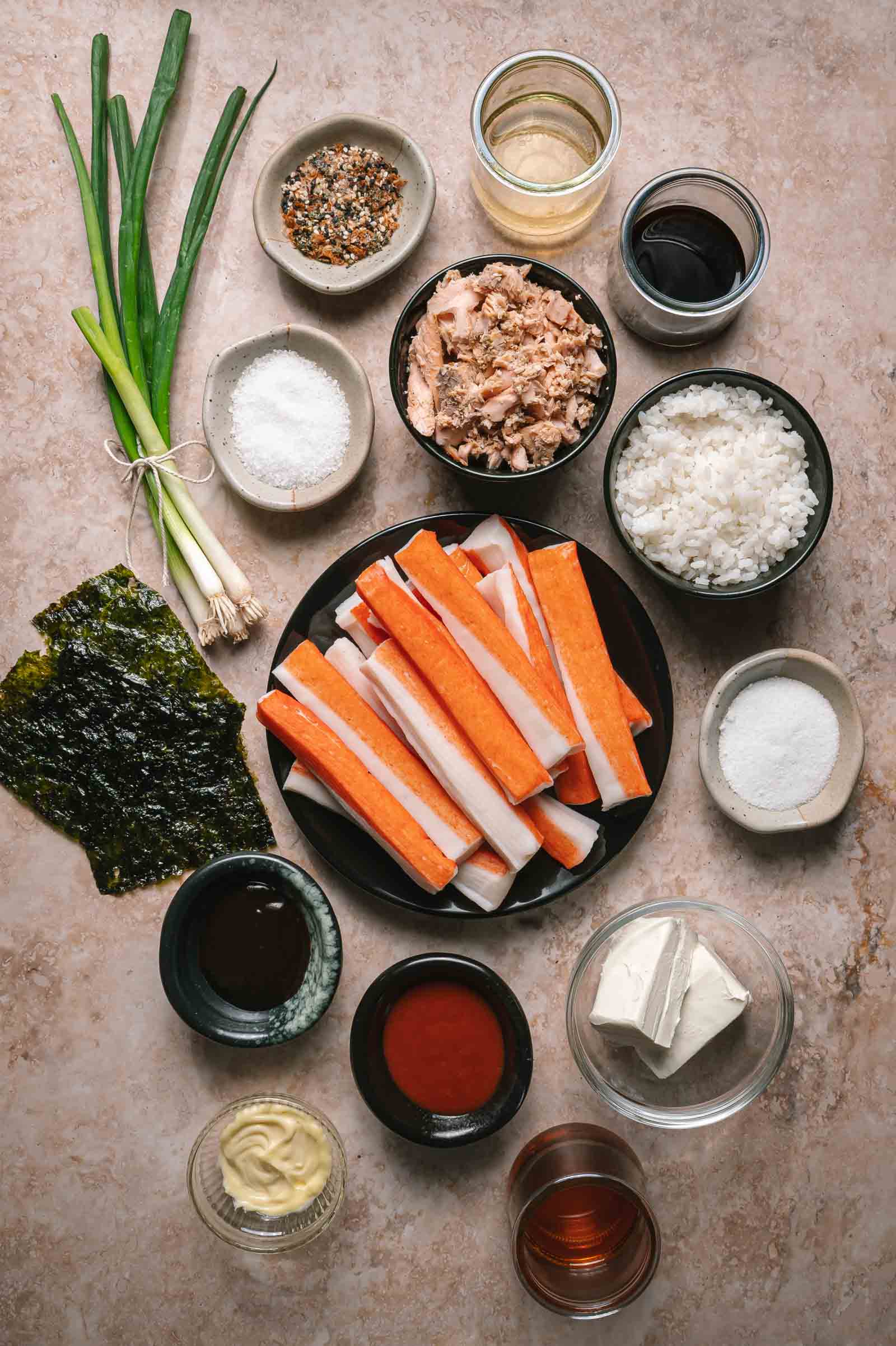 Various sushi bake ingredients, including imitation crab sticks, rice, nori, green onions, sauces, seasonings, canned tuna, tofu, and condiments, arranged on a beige surface.