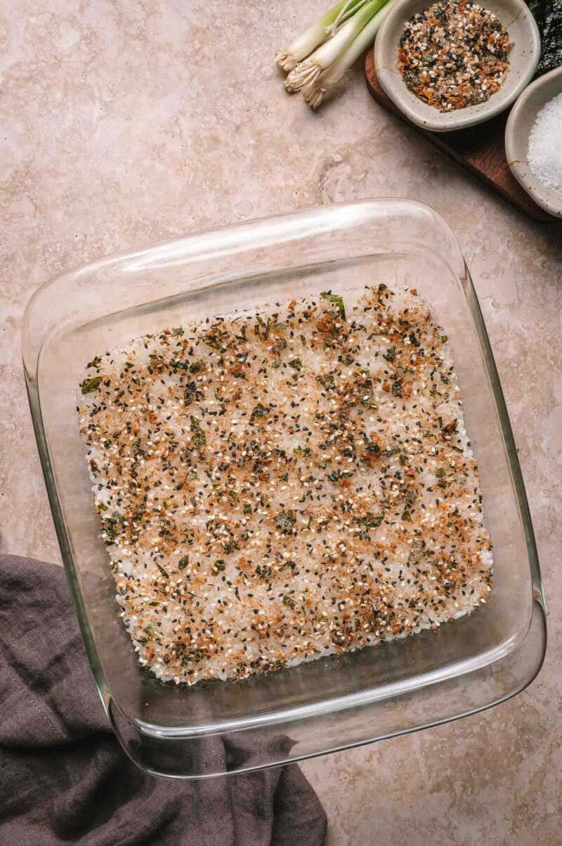 A glass baking dish contains rice with seasoning visible on top. Green onions, seasoning, and salt are in small bowls nearby.