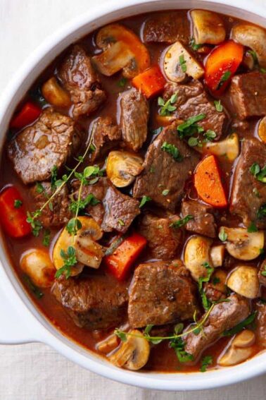 A white bowl filled with beef stew containing chunks of beef, sliced mushrooms, carrots, and garnished with chopped herbs. A spoon is placed beside the bowl.