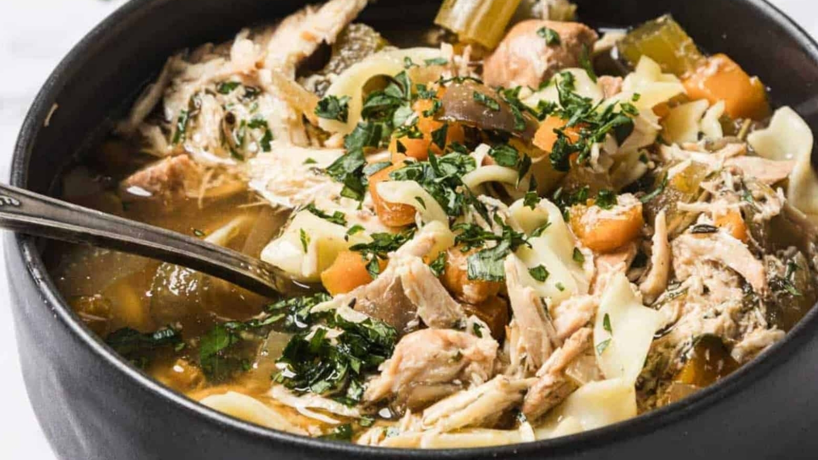 A bowl of chicken noodle soup with carrots, celery, herbs, and a spoon inside.