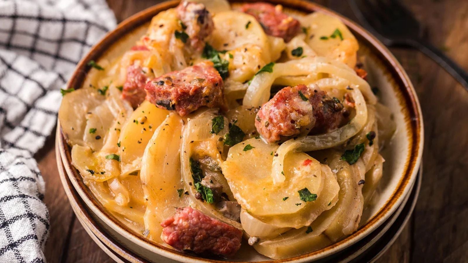 A bowl of sliced potatoes, onions, and sausage in a creamy sauce, garnished with chopped herbs.