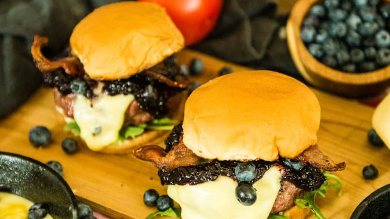 Two cheeseburgers with bacon, melted cheese, greens, blueberry sauce, and whole blueberries on buns, served on a wooden board with extra blueberries and a tomato nearby.