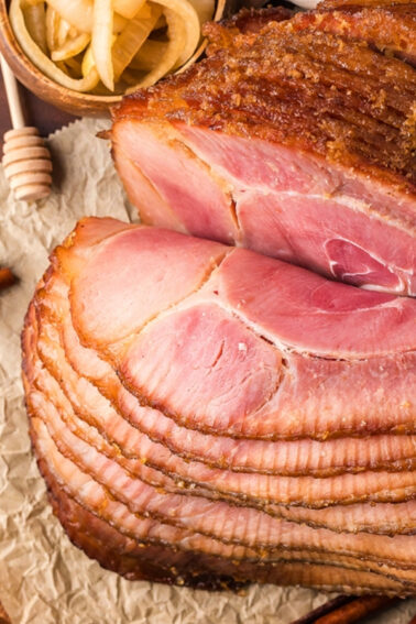 Sliced glazed ham on parchment paper atop a wooden cutting board, with a small bowl of pickled onions beside it.