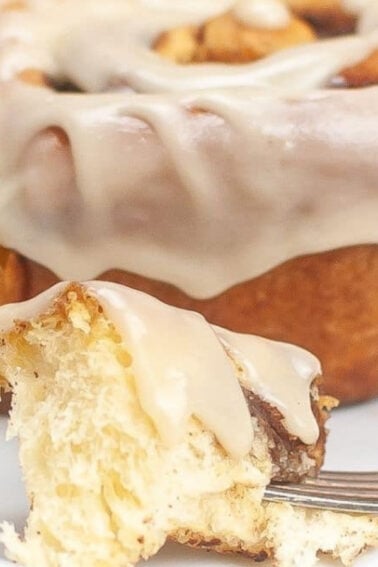 A cinnamon roll with icing sits on a white plate, with a fork holding a bite-sized piece in the foreground.
