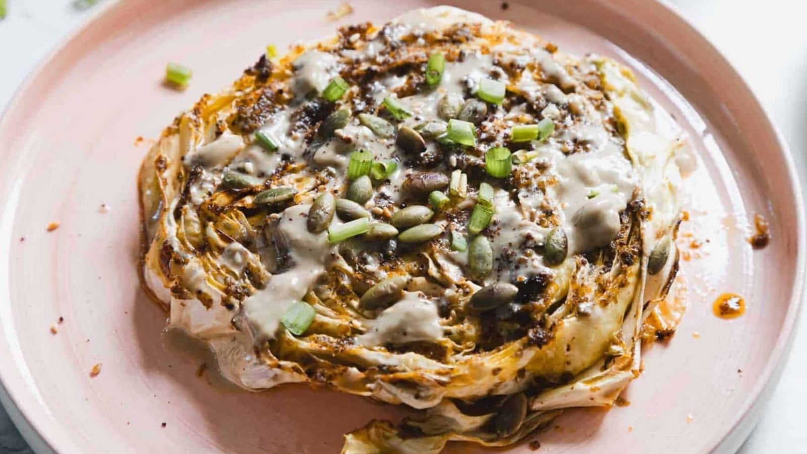 A roasted cabbage steak is served on a pink plate. It is topped with a creamy sauce, pumpkin seeds, and chopped green onions. The cabbage is browned and slightly charred at the edges.