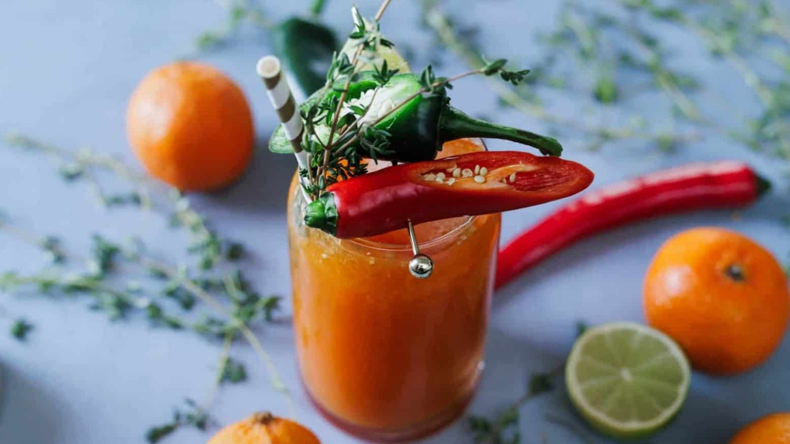 A glass filled with an orange beverage garnished with sliced chili peppers, thyme, and a straw. Surrounding the glass are whole oranges, a half lime, additional peppers, and sprigs of thyme on a light blue surface.