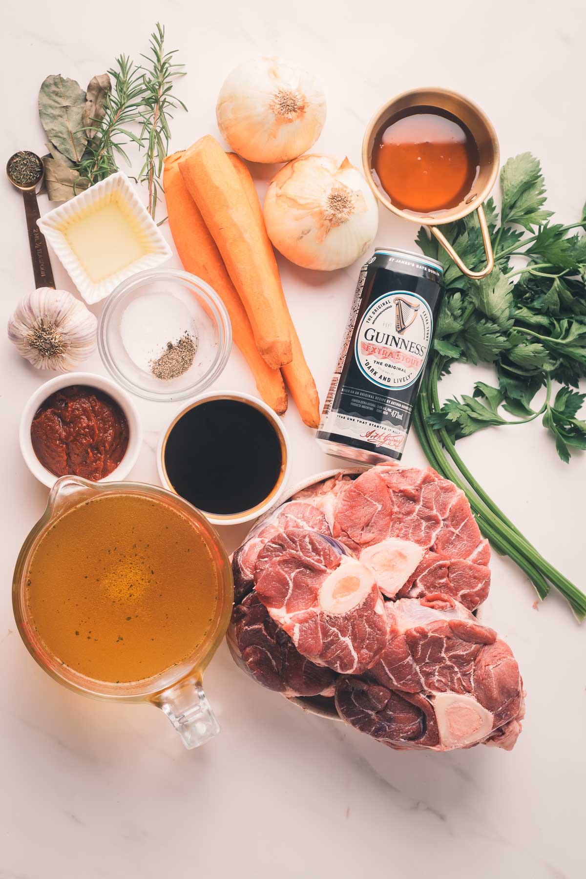 Raw ingredients for a stew, including beef shank, carrots, onions, garlic, parsley, green onions, broth, Guinness beer, tomato paste, Worcestershire sauce, seasoning, and herbs on a white surface.