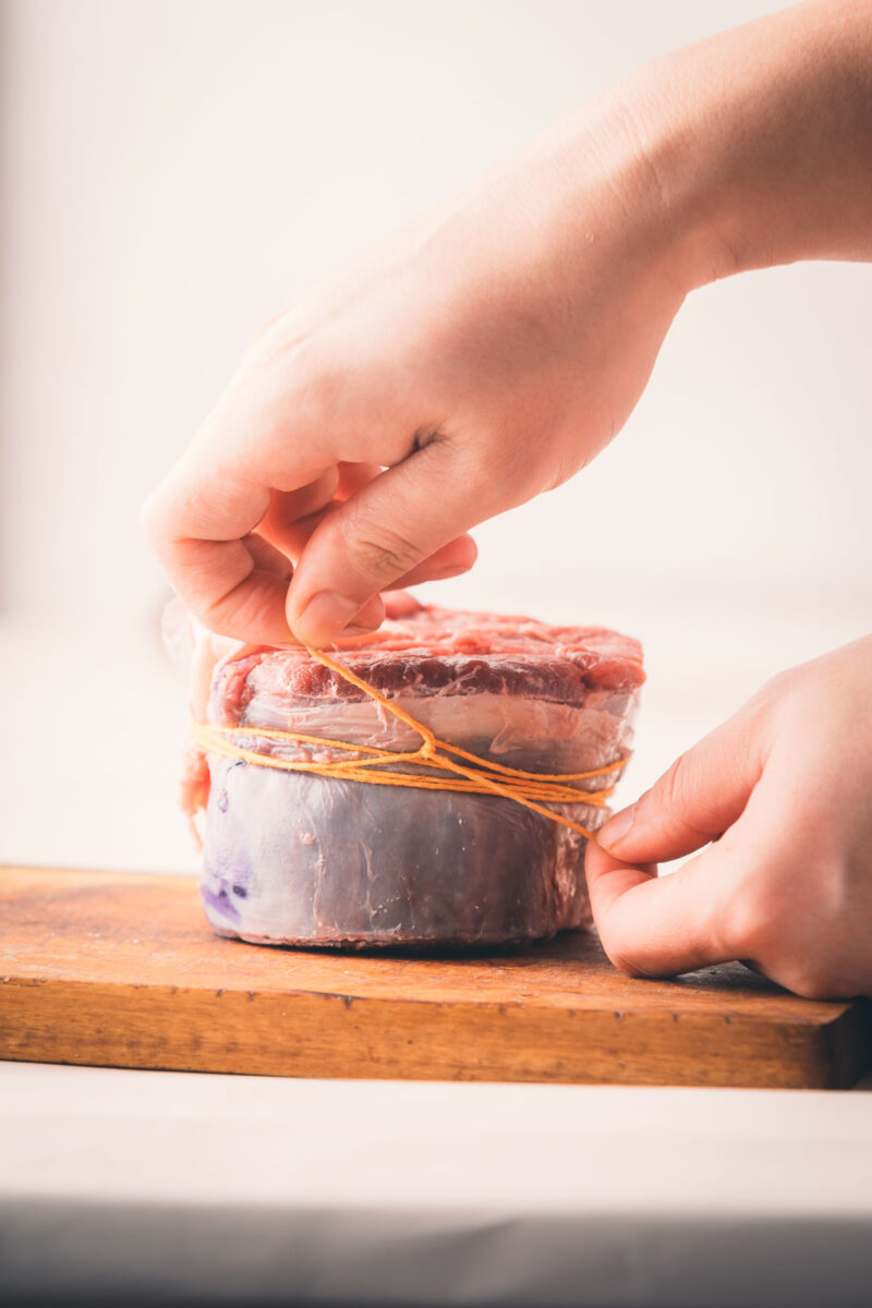A person tying kitchen twine around a beef shank on a wooden cutting board.