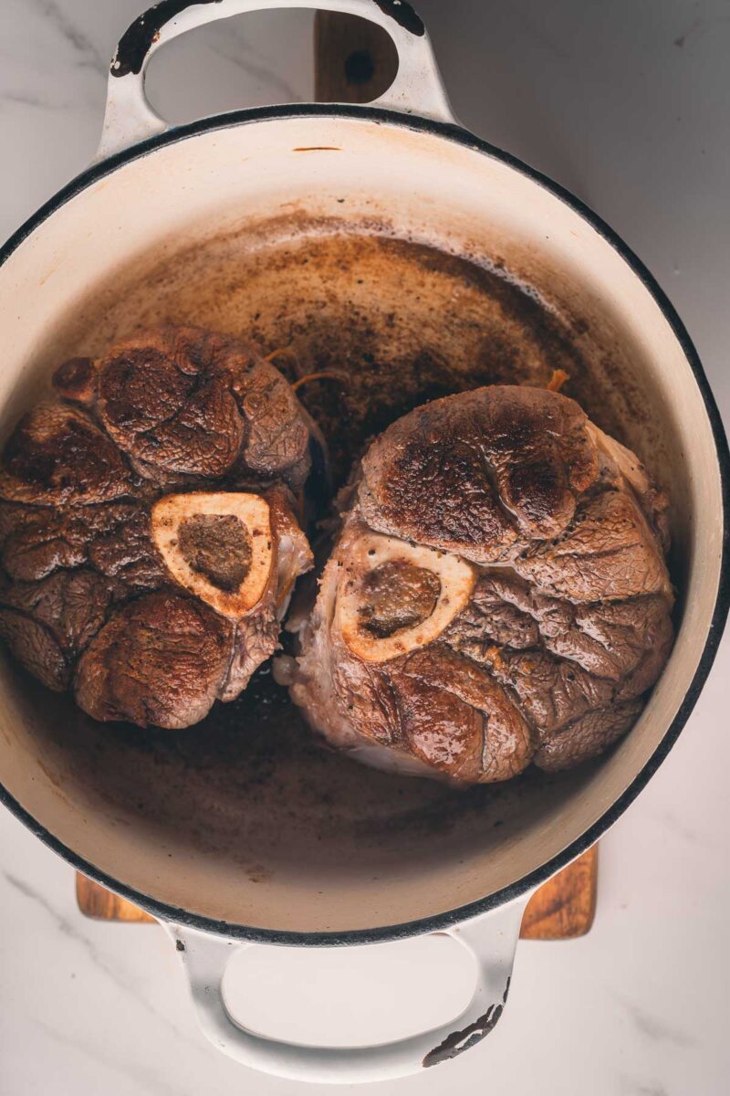 Two browned pieces of meat with bones in the center are searing in a large, round, white Dutch oven on a stovetop.