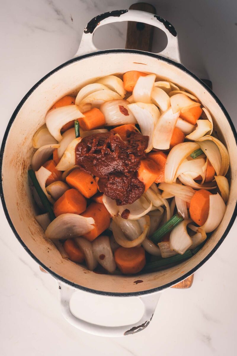Large pot containing chopped onions, carrots, celery, and a dollop of tomato paste on top, all set on a white surface.