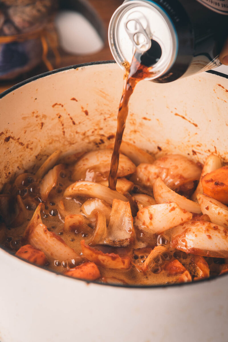 A hand pours stout beer from a can into a pot containing chopped onions, carrots, and sauce.