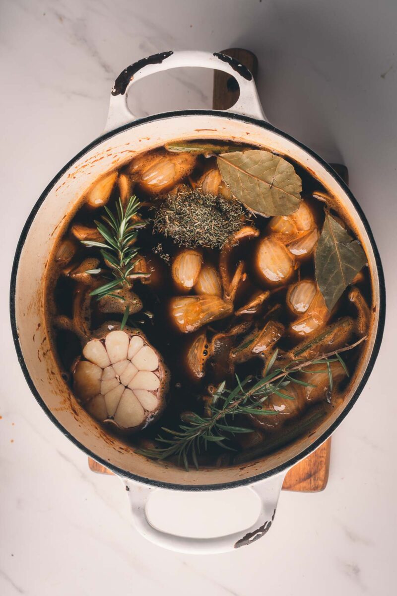 Overhead view of a pot filled with onions, garlic, bay leaves, rosemary, dried herbs, and broth on a wooden trivet, set on a white countertop.