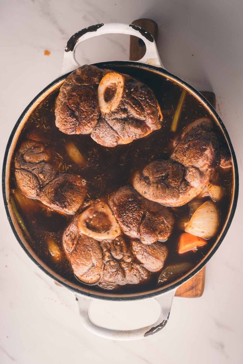 A white Dutch oven filled with braised beef shanks, vegetables, and dark broth sits on a marble surface.