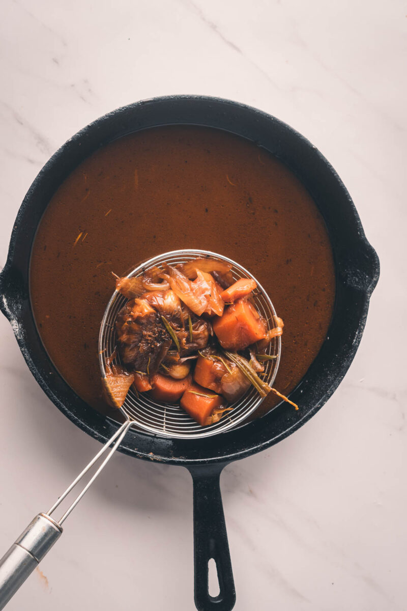 A black skillet with brown sauce and a strainer holding cooked vegetables and herbs above the liquid, on a white marble surface.