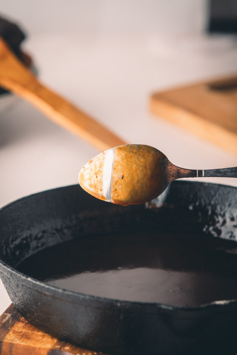 A spoon coated with the back of a brown sauce is held above a black cast iron skillet, showing the sauce’s consistency.