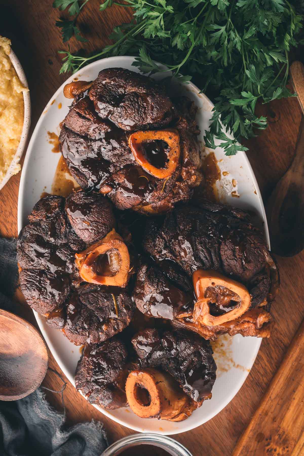 A white oval plate with three braised beef shanks, each showing a bone with marrow, placed next to fresh parsley and mashed potatoes.