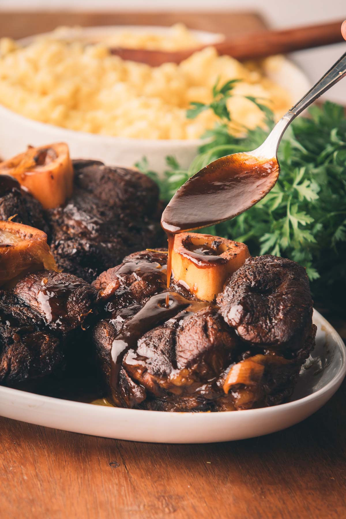 Braised beef shanks with bone marrow are served on a white plate as sauce is drizzled over them; a bowl of mashed potatoes is in the background.